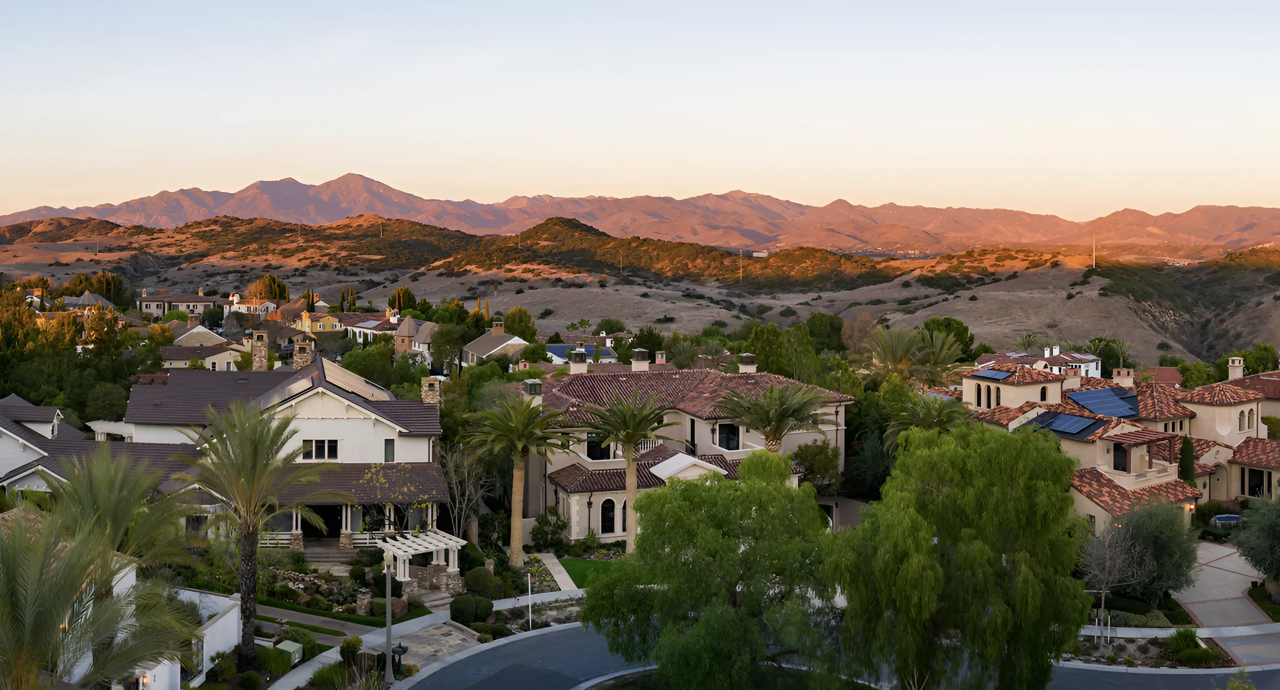 Golden-hour aerial view of fancy houses in Ladera Ranch, California, with red roofs, solar panels, and hills in the background.