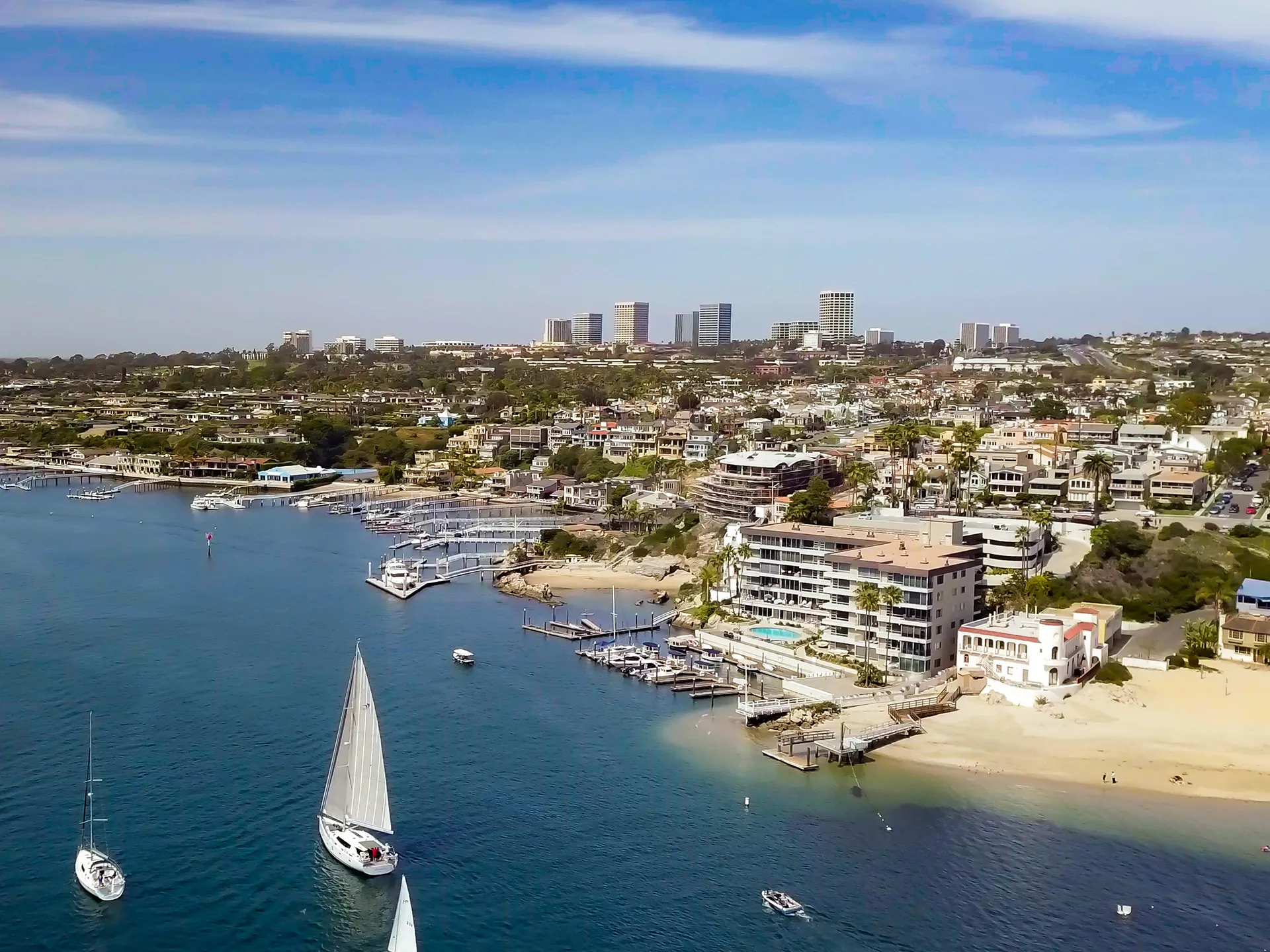 High aerial view of Corona Del Mar, Newport Beach, California, with a white sailboat, luxury beachfront homes, and Newport Center skyline in the distance.