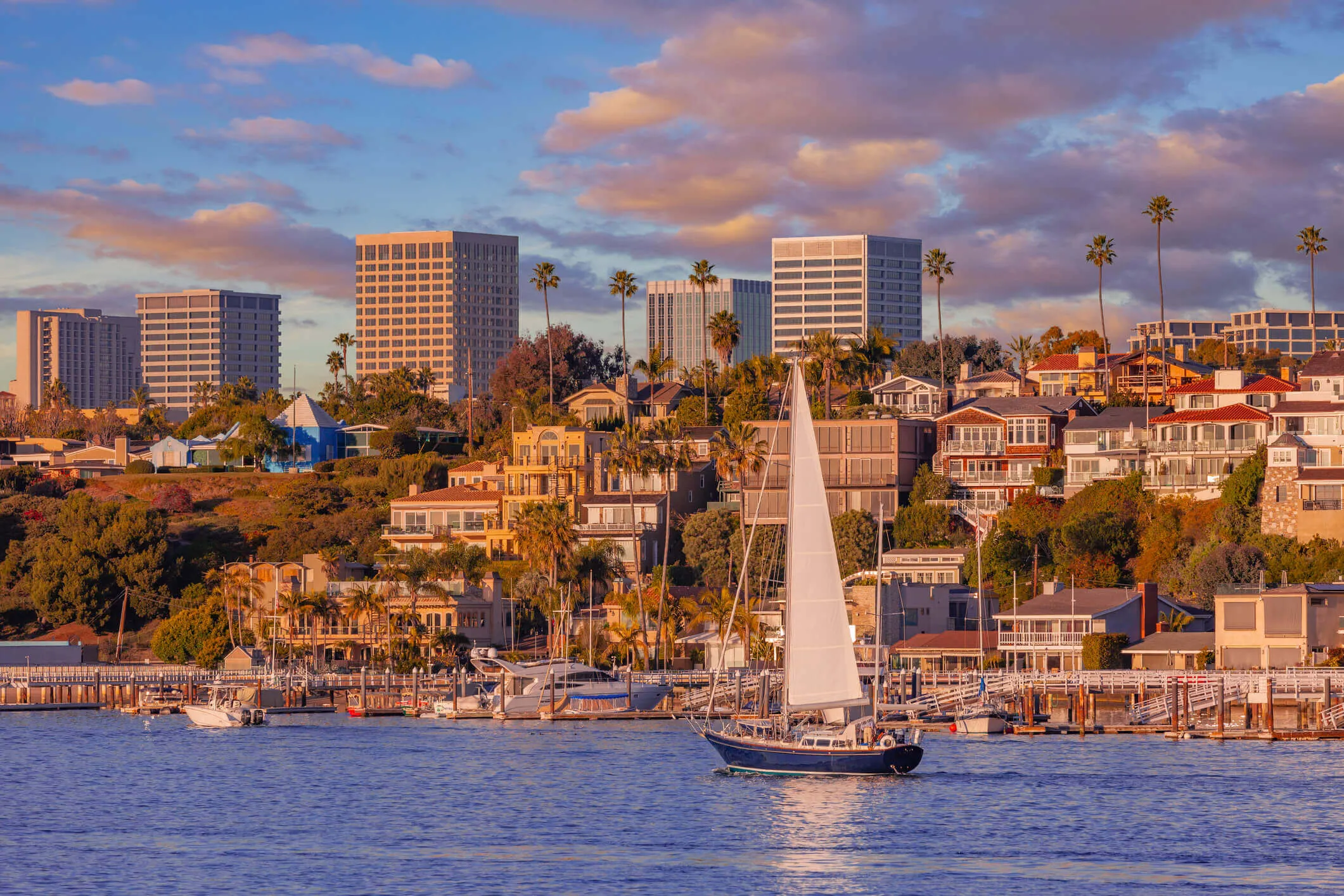 Sunset view of Newport Harbor, California, with a white sailboat, waterfront homes, and Costa Mesa skyline in the background.