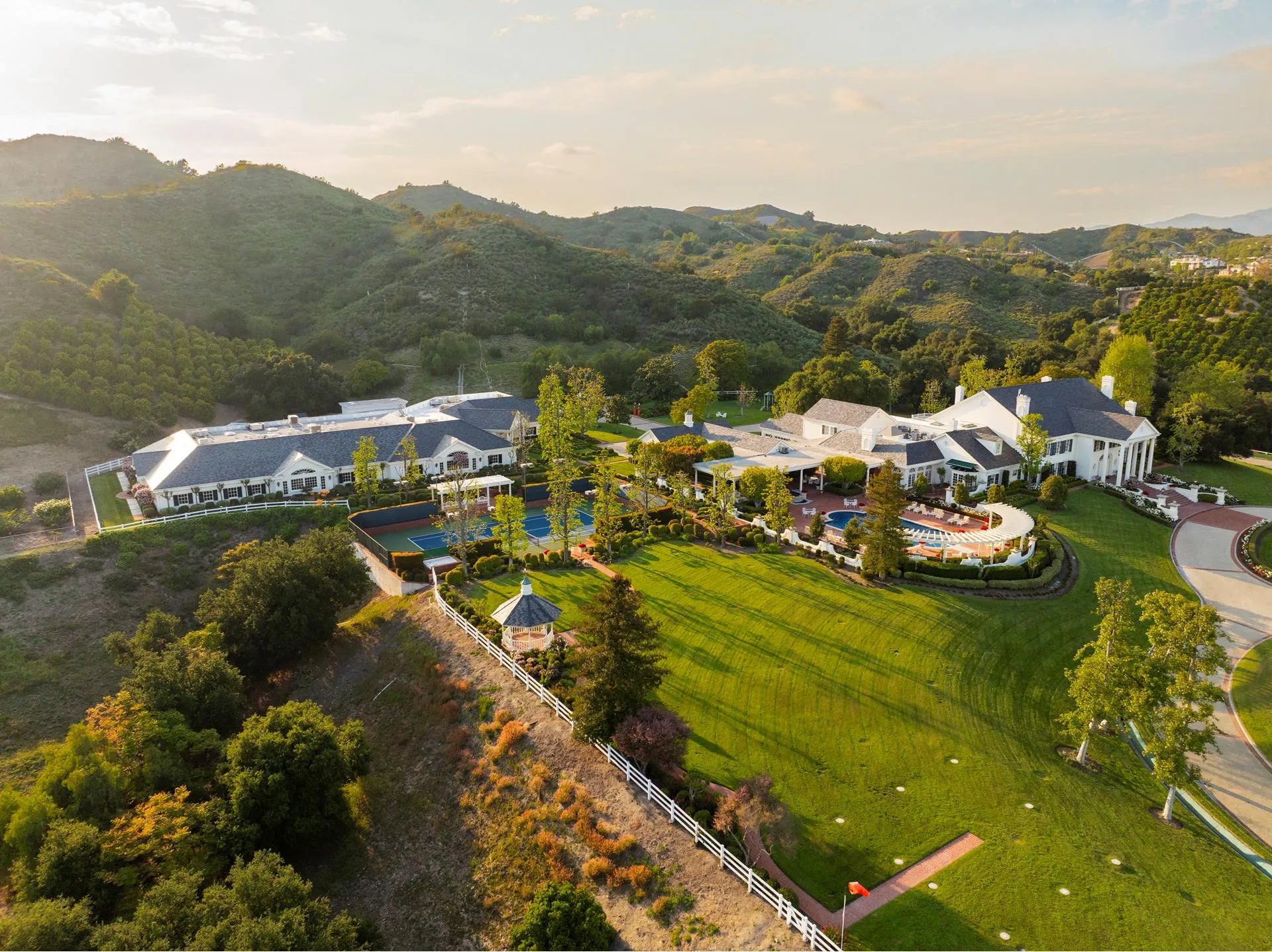 Aerial view of a luxury estate in Coto de Caza, California, with a big white house, tennis courts, pool with pergola, and green lawns by the hills.