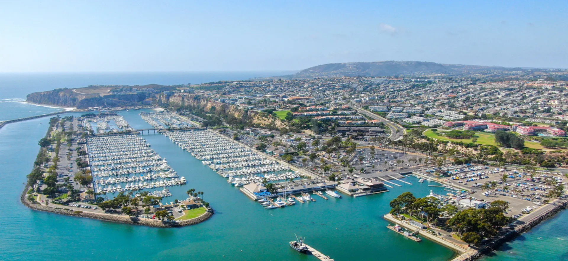 Aerial view of Dana Point Harbor, California, showing marinas with yachts, waterfront buildings, and cliffs by the ocean.