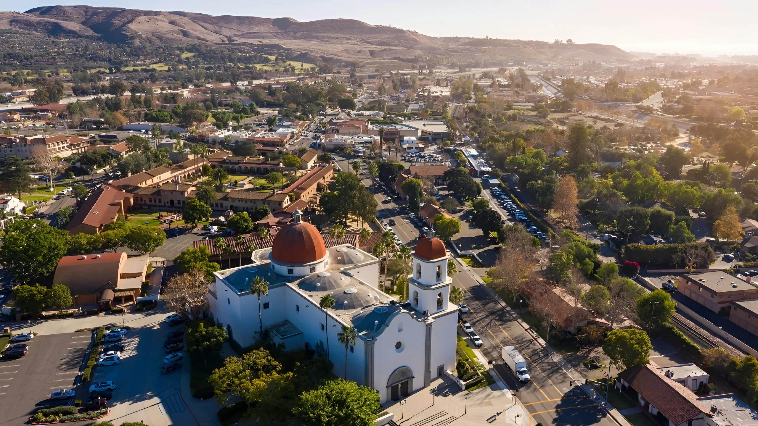 Aerial view of San Juan Capistrano, California, showing the Mission’s domed buildings and bell tower with hills in the background.